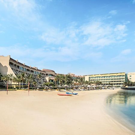 Al Raha Beach Resort & Spa Абу Даби Екстериор снимка The photo depicts a serene beach scene with a clear blue sky. In the foreground, there is a sandy beach that gently slopes down to calm water. On the left side, several palm trees provide a tropical atmosphere. In the background, there are two large 