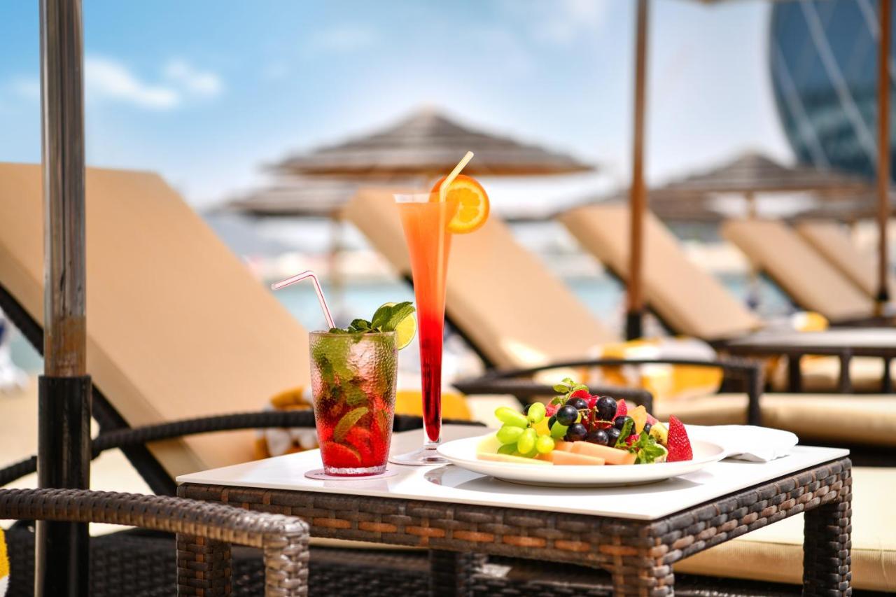 Al Raha Beach Resort & Spa Абу Даби Екстериор снимка The photo shows a table set outdoors, featuring two colorful cocktails and a plate of food. One cocktail is a vibrant red drink garnished with mint, while the other is a layered orange beverage with an orange slice on the rim. The plate of food inclu