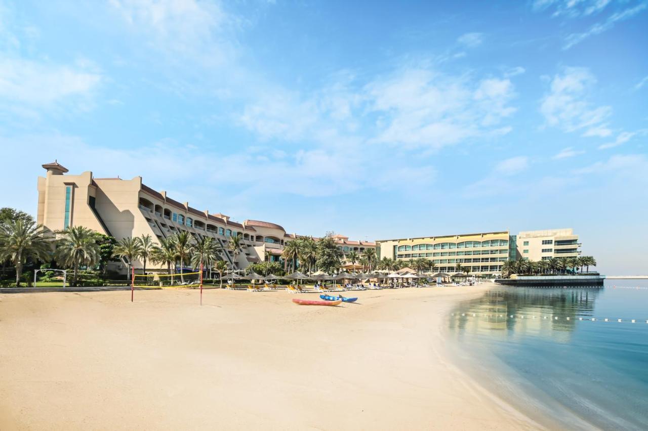 Al Raha Beach Resort & Spa Абу Даби Екстериор снимка The photo depicts a serene beach scene with a clear blue sky. In the foreground, there is a sandy beach that gently slopes down to calm water. On the left side, several palm trees provide a tropical atmosphere. In the background, there are two large 