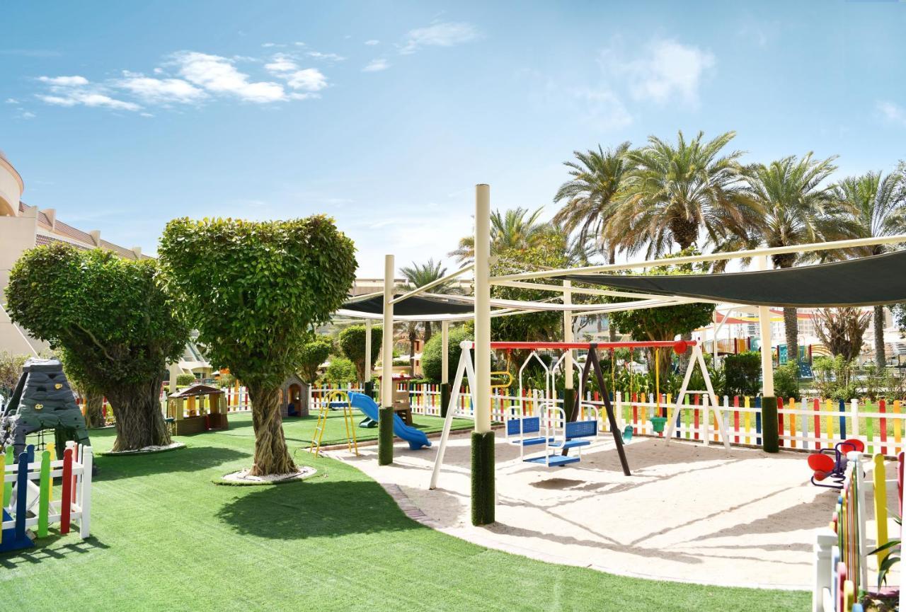 Al Raha Beach Resort & Spa Абу Даби Екстериор снимка The photo shows a brightly lit outdoor playground surrounded by palm trees. The playground features a variety of play equipment, including swings, a slide, and a climbing structure. The ground is covered in lush green artificial grass, providing a so