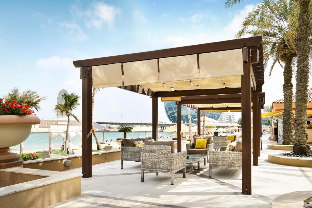 Al Raha Beach Resort & Spa Абу Даби Екстериор снимка The photo shows a stylish outdoor seating area by a beach. There is a wooden pergola covered with a light fabric shade, providing shelter to a set of comfortable wicker chairs and tables. The area is adorned with palm trees, and in the background, th