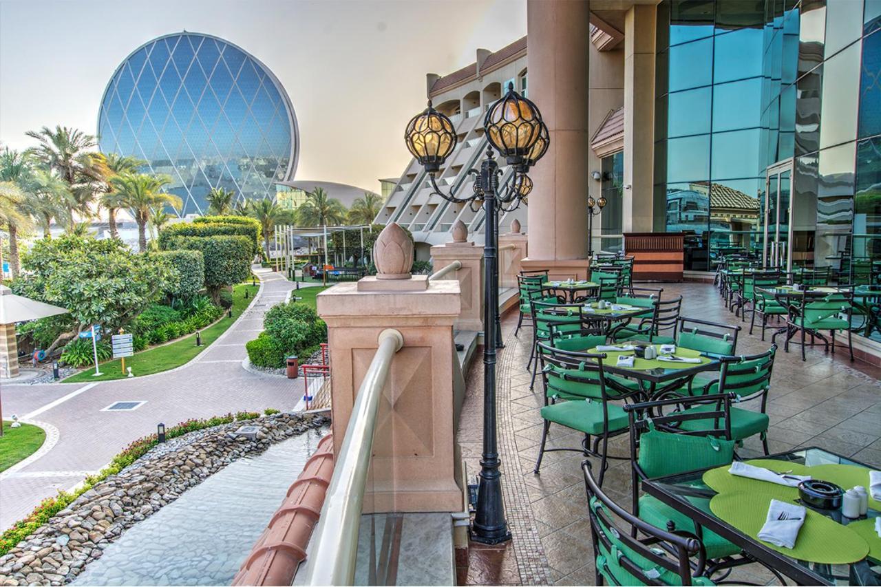 Al Raha Beach Resort & Spa Абу Даби Екстериор снимка The photo depicts an outdoor dining area with green tables and chairs arranged on a patio. In the background, there is a large, distinctive building with a striking blue dome, surrounded by well-maintained landscaping, including trees and plants. The