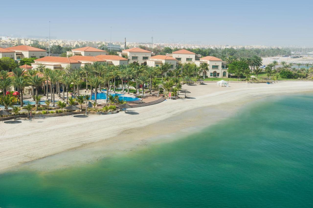 Al Raha Beach Resort & Spa Абу Даби Екстериор снимка The photo depicts a serene beach scene with a clear turquoise sea and a sandy shoreline. In the background, there are several buildings featuring a Mediterranean architectural style with red-tiled roofs. The area is adorned with palm trees and outdoo