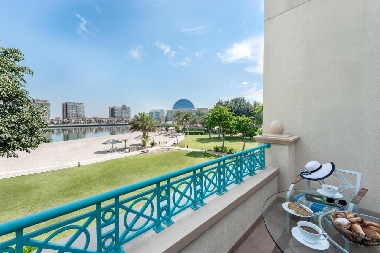 Al Raha Beach Resort & Spa Абу Даби Екстериор снимка The photo shows a balcony view overlooking a scenic landscape. In the foreground, there is a glass table set with snacks or pastries and two cups. Beyond the balcony railing, there is a well-maintained green area with palm trees and a pathway. The ba