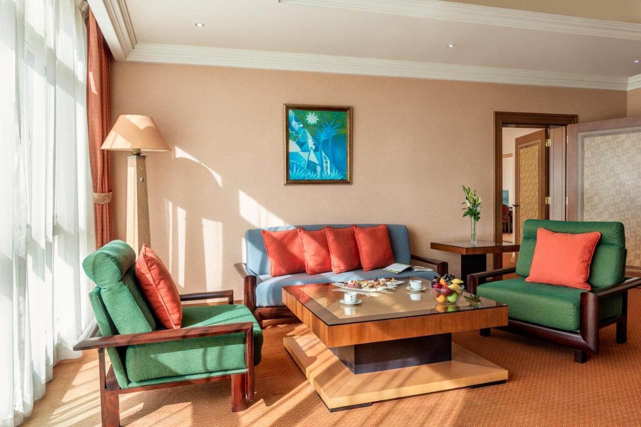 Al Raha Beach Resort & Spa Абу Даби Екстериор снимка The photo shows a cozy living room with a warm ambiance. There are two sofas: one in green with red cushions and another in blue with red cushions, arranged around a wooden coffee table. On the table, there are some fruits and a small decorative item