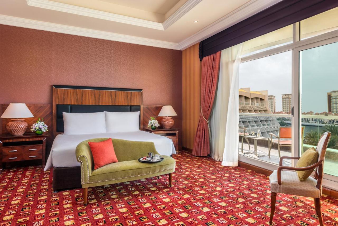 Al Raha Beach Resort & Spa Абу Даби Екстериор снимка The photo shows a hotel room with an elegant and modern design. It features a large bed with white linens and a dark wooden headboard. On either side of the bed are matching bedside lamps. There is a green armchair with a red pillow positioned near t