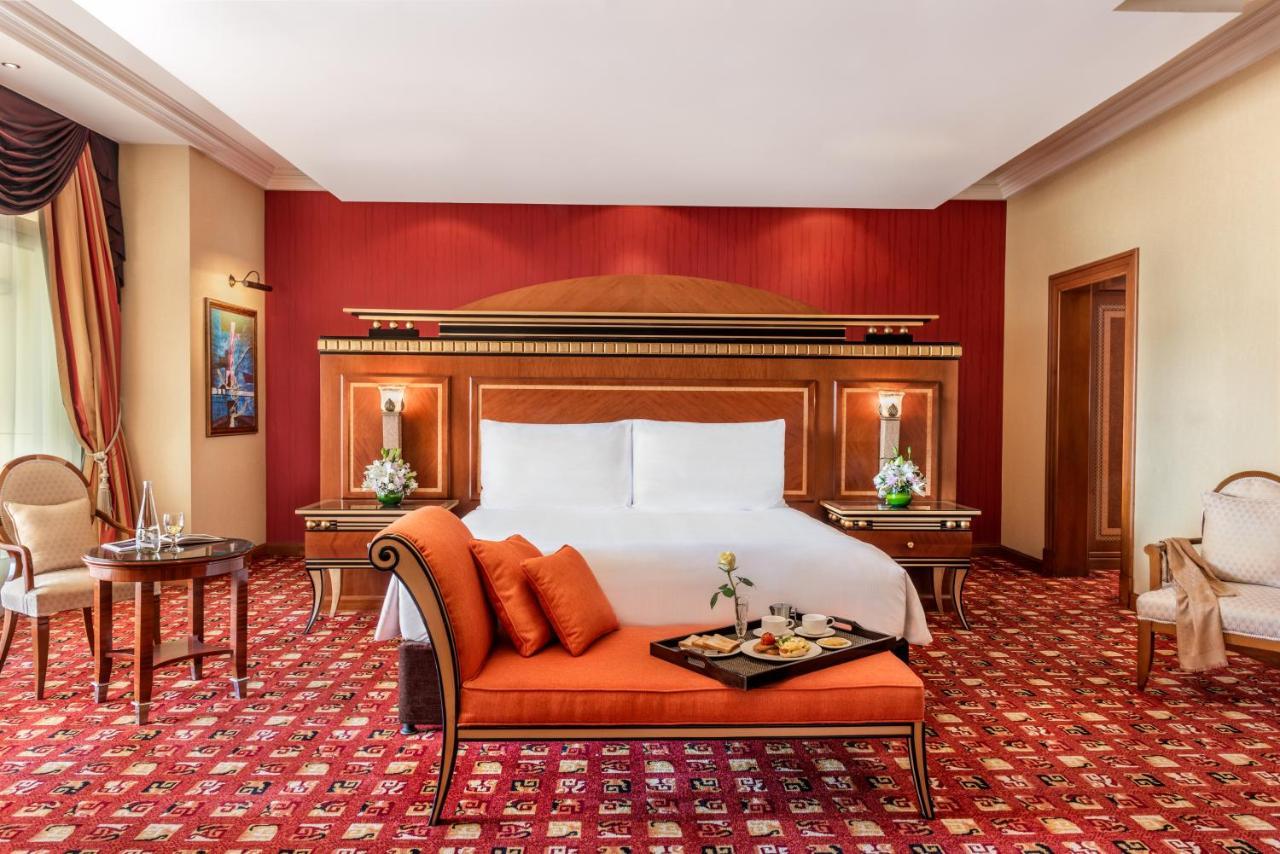 Al Raha Beach Resort & Spa Абу Даби Екстериор снимка The photo shows an elegantly designed hotel room. The room features a large bed with white linens and a decorative headboard. The walls are painted in a warm red color, and there are side tables with lamps beside the bed. 

There is an orange sofa or