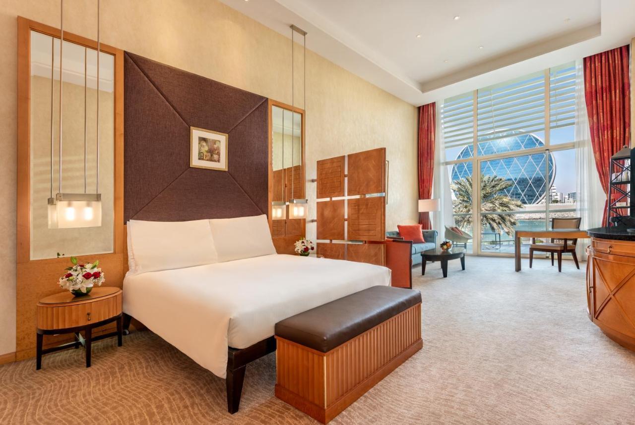 Al Raha Beach Resort & Spa Абу Даби Екстериор снимка The photo shows a modern hotel room featuring a neatly made bed with a white comforter, situated against a stylish headboard with a geometric design. There are bedside tables on either side of the bed, each with a small flower arrangement. A comforta