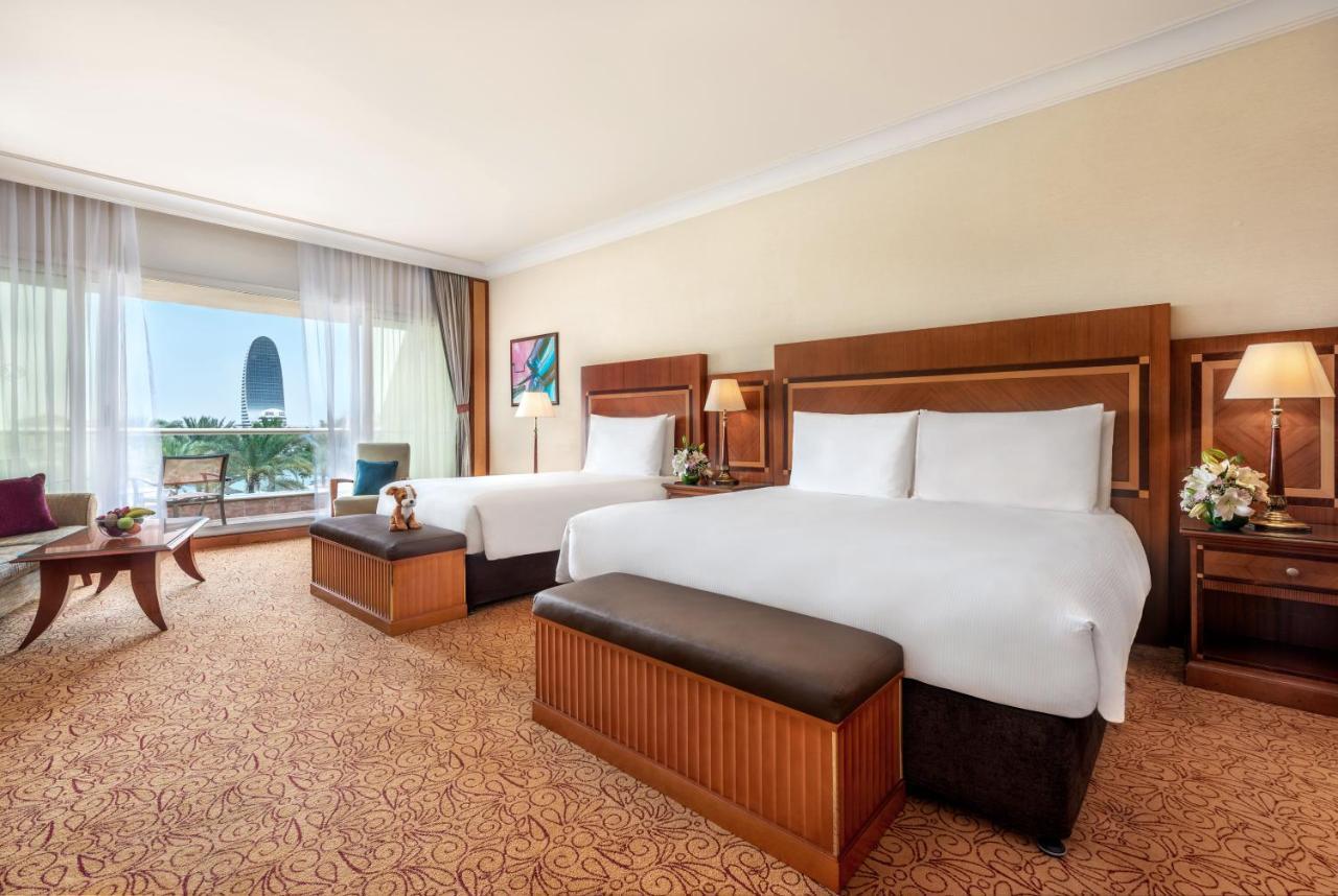 Al Raha Beach Resort & Spa Абу Даби Екстериор снимка The photo shows a spacious hotel room featuring two large beds with white linens and dark brown upholstered headboards. There are matching benches at the foot of each bed. The room is decorated with a warm color palette and has a carpeted floor. To o