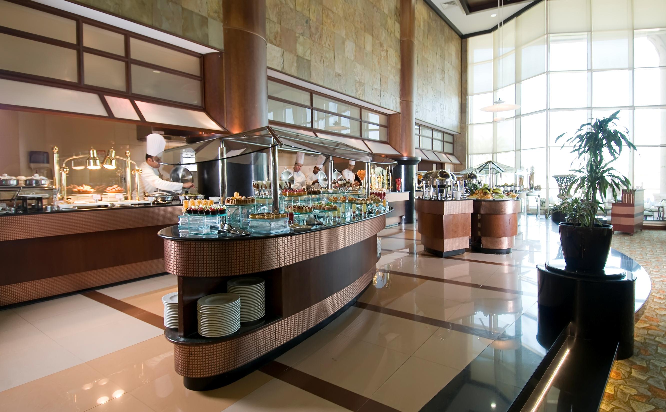 Al Raha Beach Resort & Spa Абу Даби Екстериор снимка The photo shows a spacious buffet area in a restaurant or hotel. There are multiple food stations with glass displays showcasing a variety of dishes. Chefs can be seen in the background, preparing food. The decor features large windows allowing natur