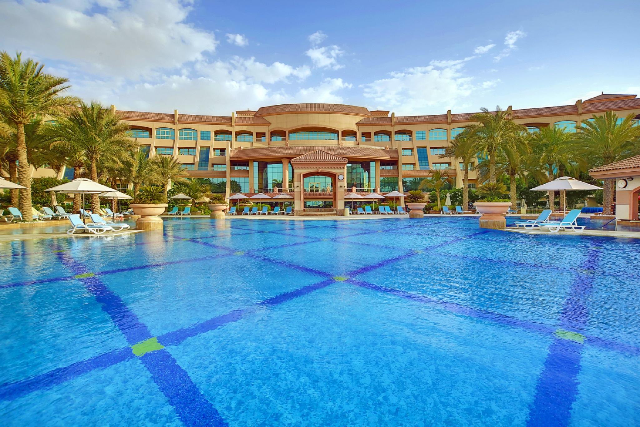 Al Raha Beach Resort & Spa Абу Даби Съоръжения снимка The photo features a luxurious resort with a large swimming pool in the foreground. The pool is crystal clear and reflects the sky, bordered by lounge chairs and palm trees. In the background, the resort building showcases modern architecture with la