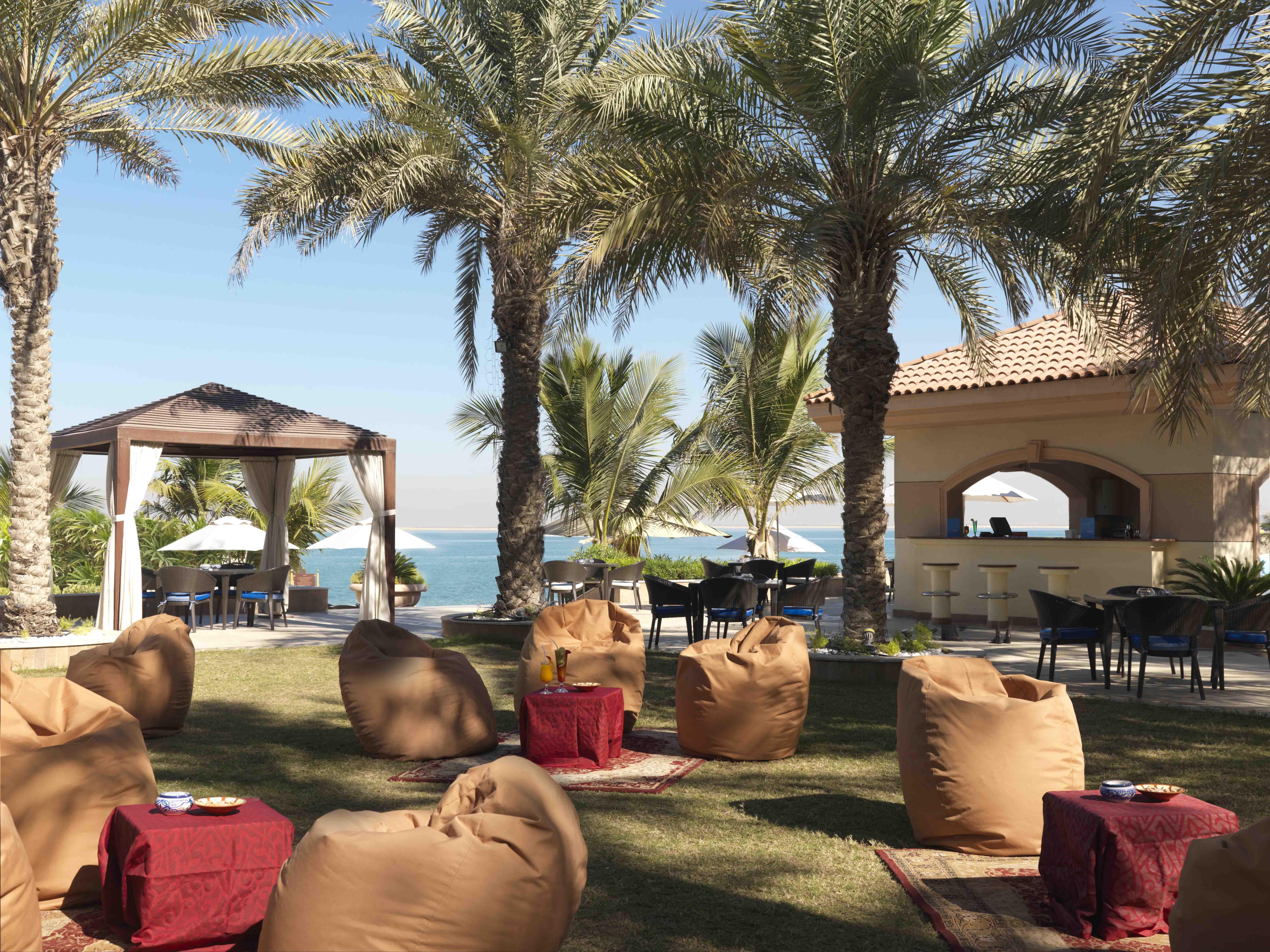 Al Raha Beach Resort & Spa Абу Даби Ресторант снимка The photo depicts a beautiful outdoor setting with several elements of relaxation and leisure. In the foreground, there are large, comfortable bean bag chairs arranged on a grassy area, accompanied by low tables. Surrounding this seating area are tal