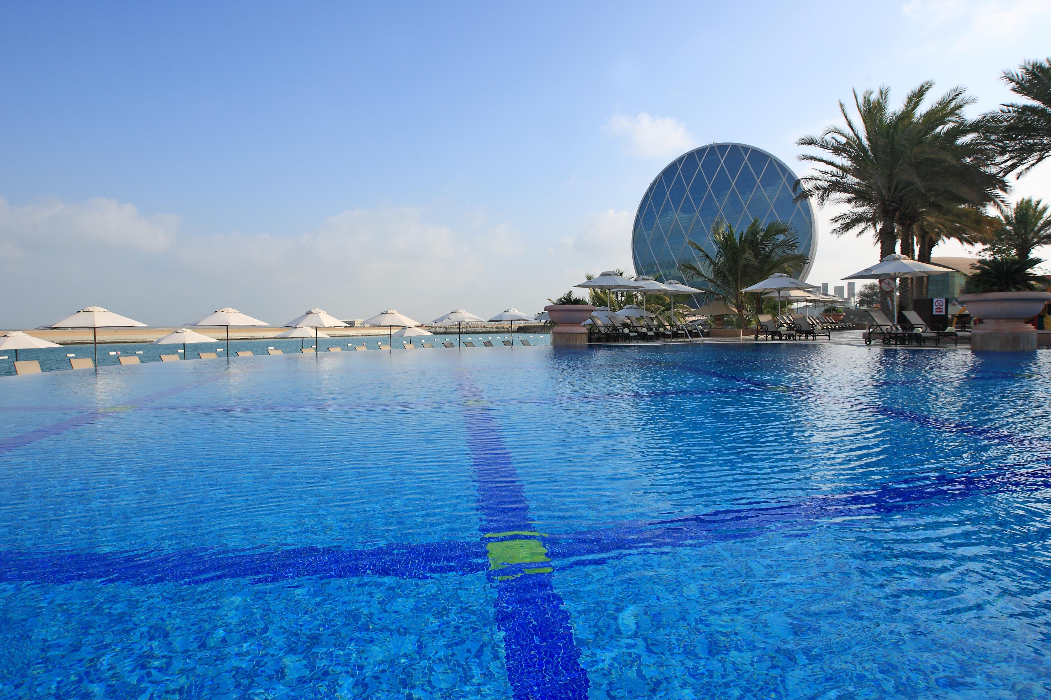 Al Raha Beach Resort & Spa Абу Даби Екстериор снимка The photo features a large, serene swimming pool with clear, blue water reflecting the sky. Surrounding the pool are several lounge chairs and umbrellas, suggesting a relaxed, resort-like atmosphere. In the background, a distinctive building with a r