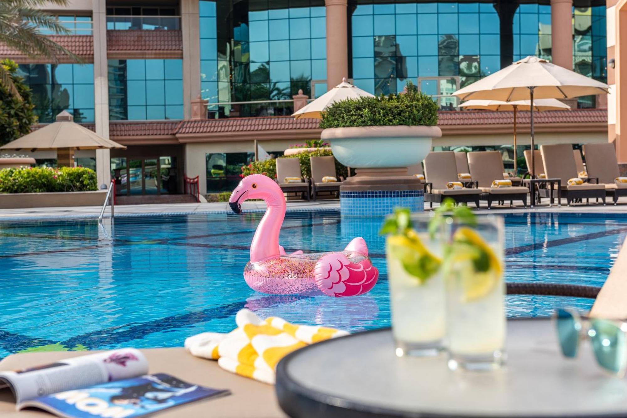 Al Raha Beach Resort & Spa Абу Даби Екстериор снимка The photo depicts a bright and inviting outdoor swimming pool surrounded by a luxurious setting. In the pool, there is a large pink flamingo float, adding a fun and whimsical touch. To the side, a table is set up with two refreshing drinks, likely co