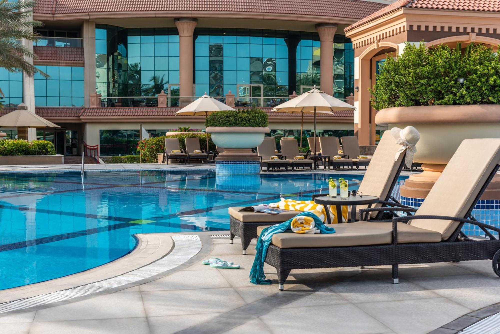 Al Raha Beach Resort & Spa Абу Даби Екстериор снимка The photo depicts a luxurious outdoor swimming pool area. In the foreground, there are two lounge chairs with cushions, positioned beside the pool. A small table between them holds drinks and snacks, suggesting a relaxing atmosphere. The pool itself 