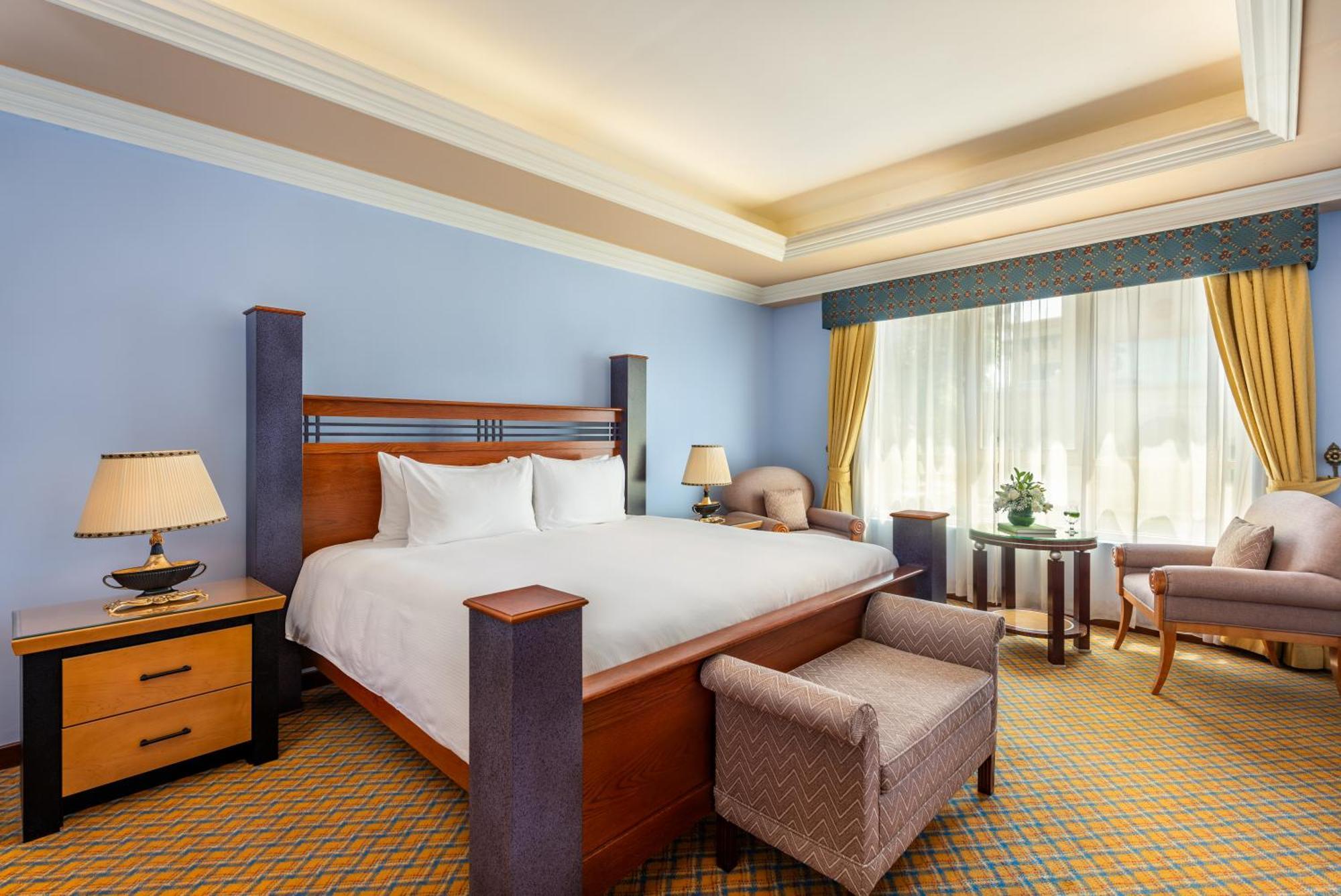 Al Raha Beach Resort & Spa Абу Даби Екстериор снимка The photo shows a well-decorated hotel room. It features a large bed with a wooden frame, neatly made with white bedding. On either side of the bed are bedside tables with lamps. There is a comfortable chair next to a window that has sheer curtains a