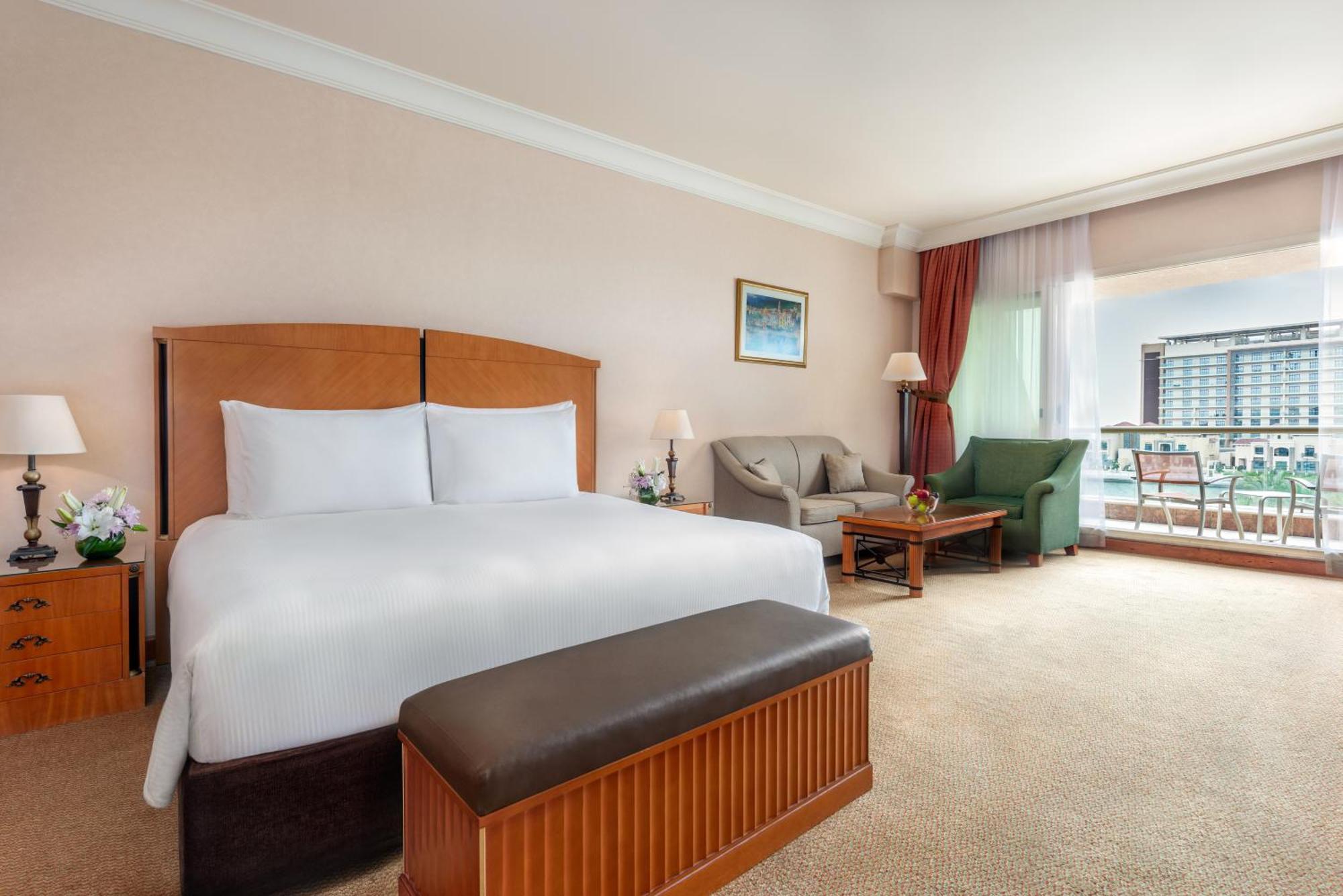 Al Raha Beach Resort & Spa Абу Даби Екстериор снимка The photo shows a well-furnished hotel room featuring a large bed with white linens and a wooden headboard. There's a brown bench at the foot of the bed. To one side, a sitting area includes a sofa and a coffee table, complemented by two lamps. Large
