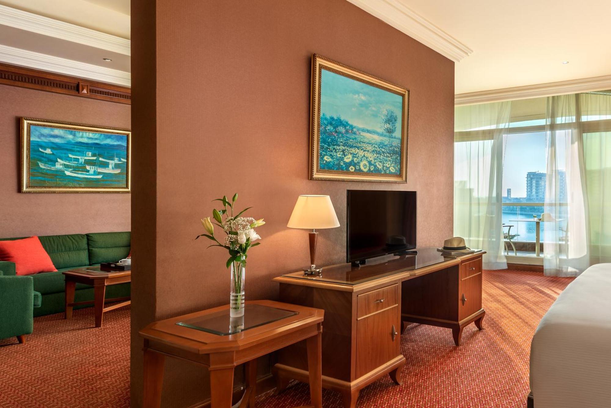 Al Raha Beach Resort & Spa Абу Даби Стая снимка The photo shows a hotel room with a warm, inviting atmosphere. There's a large window that provides natural light and a view, likely overlooking a cityscape or water. On one wall, there's a piece of artwork featuring a landscape, and nearby, a small 