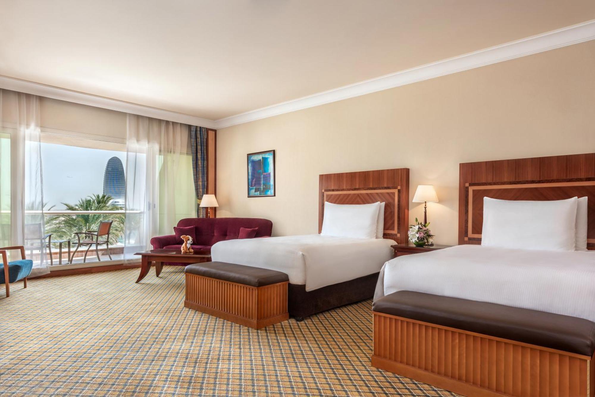 Al Raha Beach Resort & Spa Абу Даби Стая снимка The photo shows a hotel room featuring two twin beds with wooden headboards. The room has a light, neutral color scheme, with a large window that provides natural light and a view of the outdoors. There is a small seating area with a purple sofa and 