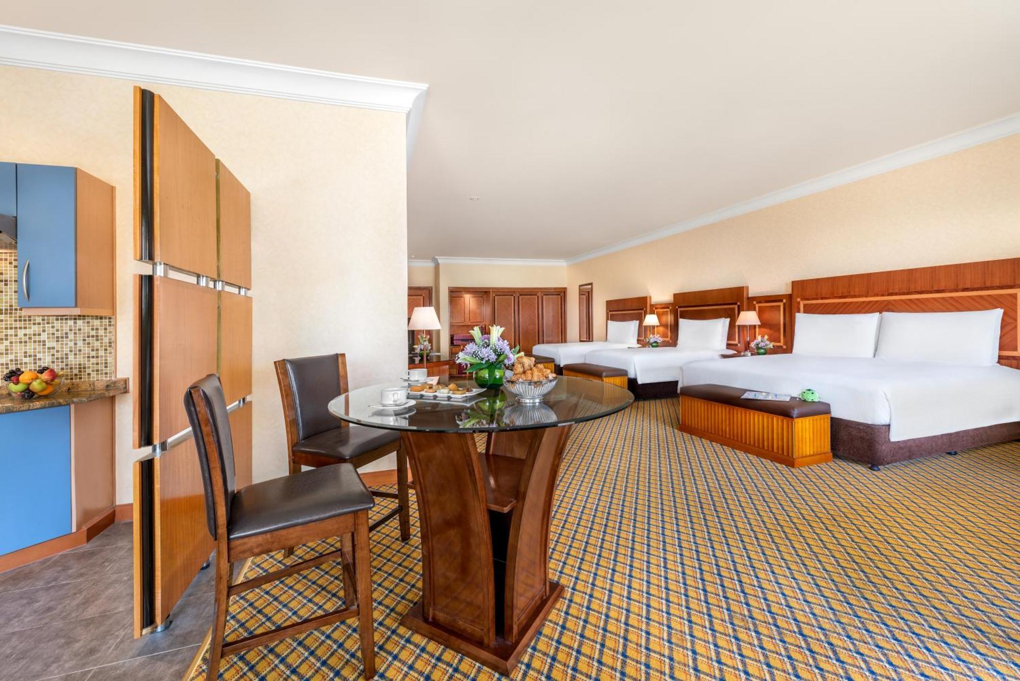 Al Raha Beach Resort & Spa Абу Даби Стая снимка The photo displays a spacious hotel room. It features two large beds with white bedding, a circular glass dining table with a decorative floral centerpiece, and a couple of chairs around it. There is a wooden cabinet along one wall, and the room has 