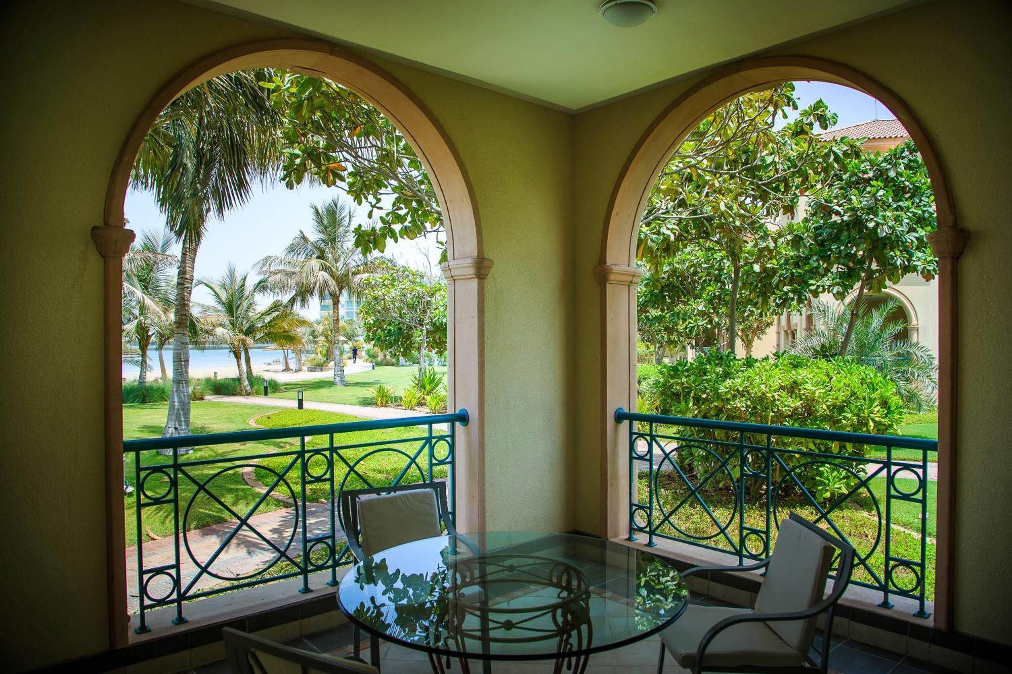Al Raha Beach Resort & Spa Абу Даби Стая снимка The photo displays a beautifully designed balcony or terrace area. It features two large arches that frame the view outside. Through the arches, you can see a lush garden with palm trees and well-maintained greenery. There’s a glass table with severa