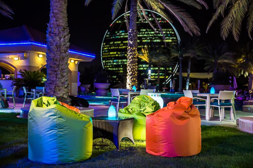 Al Raha Beach Resort & Spa Абу Даби Екстериор снимка The photo shows a vibrant outdoor lounge area at night. In the foreground, there are colorful bean bag chairs—one in bright green, one in yellow, and one in orange. A small table sits between the bean bags. In the background, illuminated palm trees a