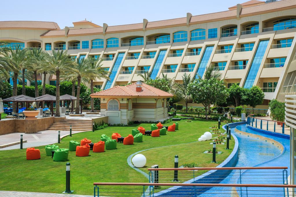 Al Raha Beach Resort & Spa Абу Даби Екстериор снимка The photo depicts a modern hotel complex with multiple stories and large windows. In the foreground, there is a well-maintained lawn featuring colorful lounge chairs in shades of orange and green. A pathway runs alongside a blue water feature, creati