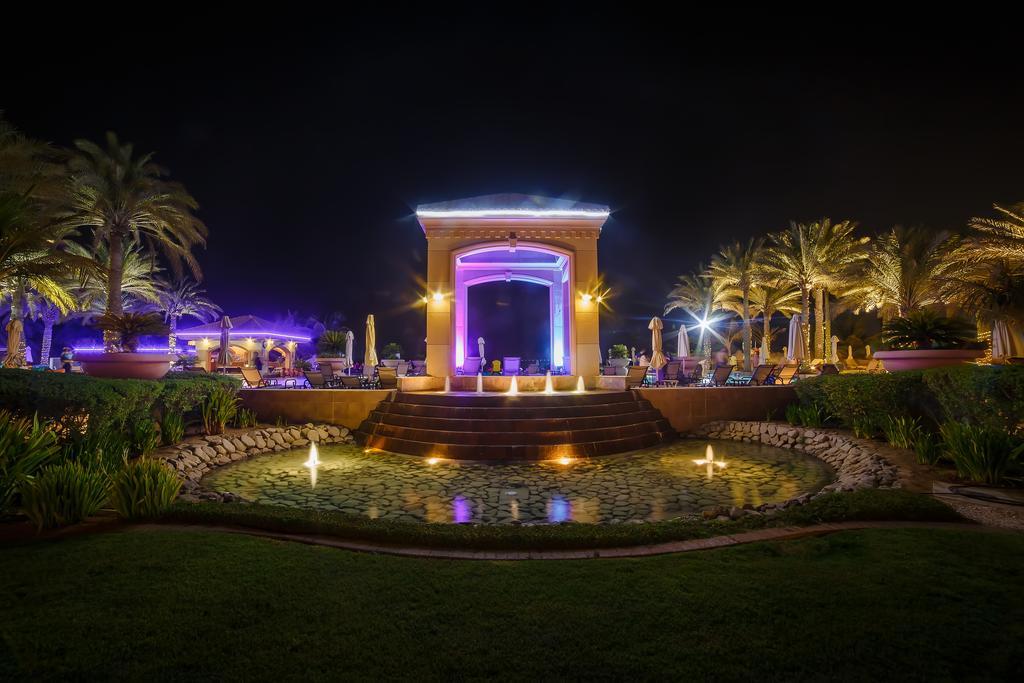 Al Raha Beach Resort & Spa Абу Даби Екстериор снимка The photo depicts a beautifully lit outdoor scene at night. In the foreground, there is a serene water feature with a circular pond, and small fountains creating gentle ripples. Surrounding the pond are lush green lawns and ornamental plants. 

In th