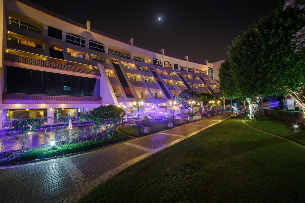 Al Raha Beach Resort & Spa Абу Даби Екстериор снимка The photo shows a modern building illuminated at night, with a vibrant purple hue. The structure has multiple floors, each with balconies overlooking a landscaped area. In the foreground, there is a pathway lined with well-manicured grass and shrubs,