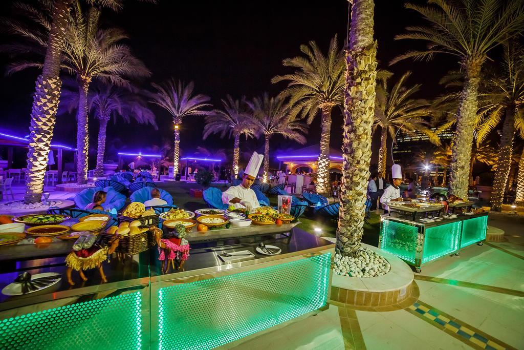 Al Raha Beach Resort & Spa Абу Даби Екстериор снимка The photo depicts an outdoor dining area at night, featuring several chefs in white uniforms and hats working at a food station. The setting is illuminated by colorful lights, with glowing green accents on the serving tables. Surrounding the area are